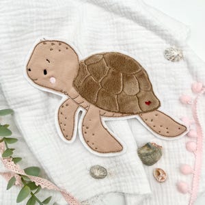 Può includere: Applicazione di tartaruga marrone e beige con bordo bianco, con guscio strutturato e un piccolo cuore rosso. L'applicazione è su un tessuto bianco strutturato, con conchiglie e finiture con pompon rosa.