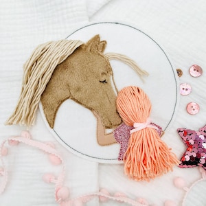May include: A brown horse with a blonde mane and a pink girl with long orange hair are embroidered on a white felt circle. The horse is nuzzling the girl's head.