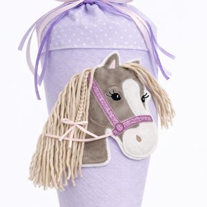 Könnte beinhalten: Ein lavendelfarbener, kegelförmiger Geschenktüte mit einem Pferde-Applikation. Das Pferd hat einen grauen Kopf, eine weiße Schnauze und einen rosa Zaum. Die Tüte ist mit lila und rosa Bändern gebunden.