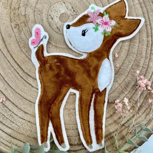 Op de afbeelding: Een bruine hertenapplicatie met een witte kop en accenten, versierd met een bloemenkrans en een roze vlinder. Het hert is gemaakt van een zacht, pluche materiaal en is geschikt voor knutselen of decoratie.