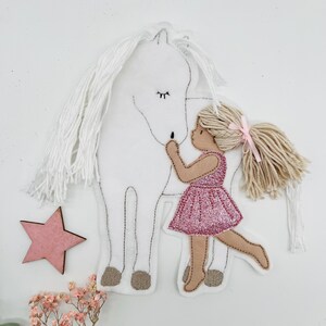 Op de afbeelding: Een vilten decoratie met een wit paard met een lange, witte manen en een meisje in een roze glitterjurk dat het paard omhelst. Een roze ster en kleine roze bloemen zijn ook inbegrepen.