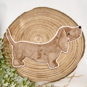 Könnte beinhalten: Ein brauner Dackel aus Plüsch mit weißer Umrandung, auf einem natürlichen Holzscheiben-Hintergrund. Der Hund hat eine schwarze Nase, rosa Wangen und einen freundlichen Ausdruck. Geeignet für Kinderzimmerdekoration.