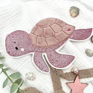 Könnte beinhalten: Ein rosa glitzernder Schildkröten-Applikation mit einem plüschigen, strukturierten Panzer. Die Schildkröte hat eine weiße Kontur, ein kleines rosa Herz und ein süßes, lächelndes Gesicht. Dekorative Muscheln und ein Stern akzentuieren den Hintergrund.