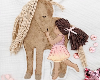 May include: A fabric applique featuring a brown horse and a girl in a pink dress. The horse has a tan mane and the girl has brown hair with a pink bow. Pink buttons and a sequined star are also present.