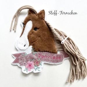 Könnte beinhalten: Ein brauner Plüschpferdekopf mit weißem Gesicht und einem rosa Band. Der Pferdekopf ist an einem weißen Stoffbanner mit rosa Glitzer und dem in rosa gestickten Text "Ponyliebe" befestigt. Die Mähne des Pferdes besteht aus beigefarbenem Garn.