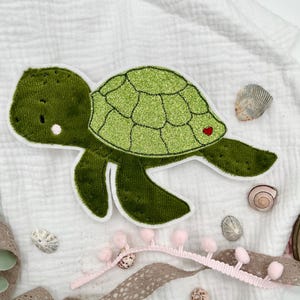 Könnte beinhalten: Eine grüne Filz-Schildkröten-Applikation mit Glitzer auf dem Panzer und einem kleinen roten Herz auf dem Panzer. Die Schildkröte befindet sich auf einem weißen Stoffhintergrund.
