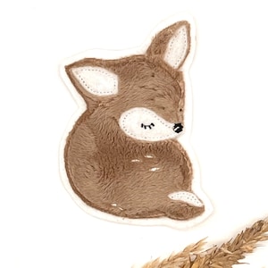 Könnte beinhalten: Ein braunes Plüsch-Rehkitz-Applik mit weißen Nähten und geschlossenen Augen. Das Rehkitz ist in einer Schlafposition zusammengerollt.