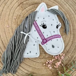 Può includere: Una testa di cavallo in feltro bianco con segni grigi a pois e una criniera grigia. Il cavallo ha una briglia viola glitterata e un piccolo fiocco bianco. L'oggetto è decorativo.