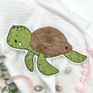 Könnte beinhalten: Eine grüne und braune Filzapplikation einer Schildkröte mit Glitzerfinish. Die Schildkröte hat eine braune Schale und einen grünen Körper mit einem weißen Gesicht.