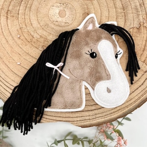 Könnte beinhalten: Ein Plüschpferdekopf mit braunem Körper, weißem Gesicht und schwarzer Mähne. Das Pferd hat eine kleine rosa Schleife und ist auf einem Holzscheiben-Hintergrund platziert. Es ist ein dekorativer Artikel.