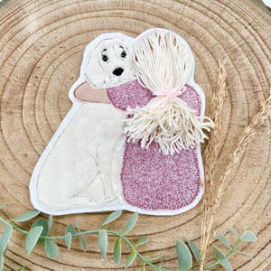 Könnte beinhalten: Ein gestickter Patch mit einem Mädchen, das einen Hund umarmt. Das Mädchen trägt ein rosa Kleid und hat lange blonde Haare. Der Hund ist weiß und hat eine schwarze Nase.
