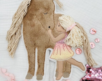 May include: A brown plush horse with a long blonde mane and tail is being hugged by a girl with blonde hair wearing a pink and yellow dress. The girl is holding a pink bow in her hair. The image is on a white background with pink and white pom-poms.
