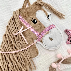 Könnte beinhalten: Ein brauner und weißer Plüschpferdekopf mit einer geflochtenen rosa Mähne und einem rosafarbenen, schimmernden Zaumzeug. Der Pferdekopf ist an einer langen, geflochtenen braunen Mähne befestigt.