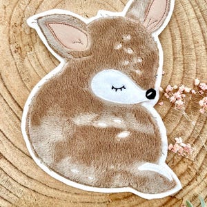 Op de afbeelding: Een bruine hert-applicatie met witte vlekken en een witte rand. Het hert heeft gesloten ogen, een zwarte neus en roze binnenoren. De applicatie is geplaatst op een houten achtergrond met een paar roze bloemen.