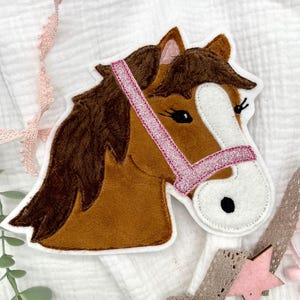 Può includere: Un'applicazione a forma di testa di cavallo marrone con un morso rosa glitterato. Il cavallo ha una faccia bianca e una pelliccia marrone. L'applicazione è perfetta per aggiungere un tocco di fantasia a un progetto artigianale.