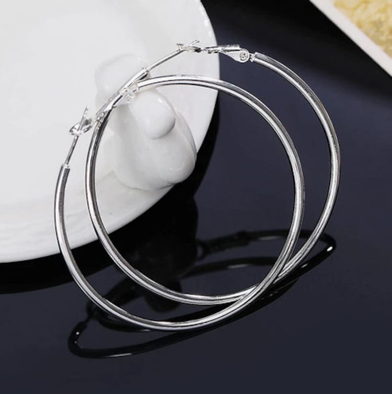 May include: Two silver-colored hoop earrings. The earrings are round and have a simple, polished design. They are resting on a white ceramic surface, with a dark background. The earrings are a classic jewelry item.