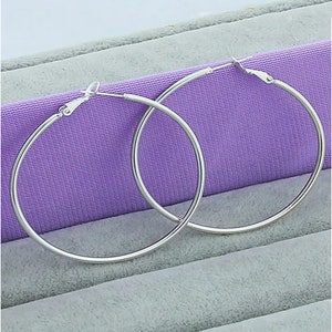 May include: A pair of large, silver-colored hoop earrings. The earrings are circular and have a simple, polished design. They are displayed on a purple surface.