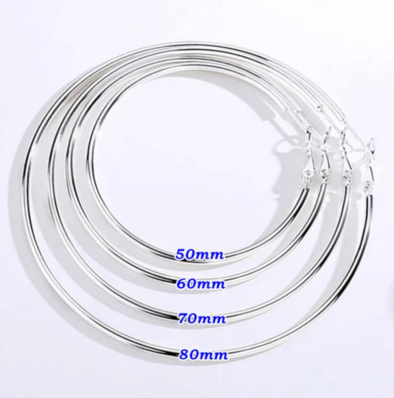 May include: Four silver hoop earrings in graduated sizes: 50mm, 60mm, 70mm, and 80mm. The earrings are displayed on a white background. Each earring has a simple, polished design with a clasp.