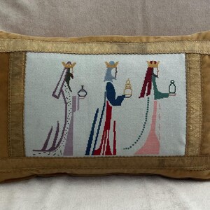 May include: A rectangular pillow with a tan velvet exterior and a central embroidered panel. The panel depicts three figures in robes, each holding an item. The figures are in shades of purple, red, blue, and green, with gold crowns.