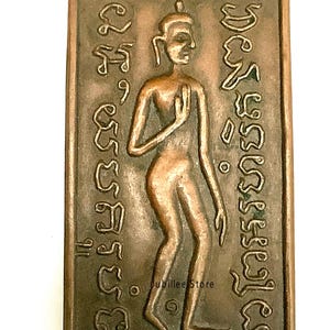 Puede incluir: Un amuleto rectangular de color bronce que representa una figura de pie con una mano levantada. La figura está rodeada de una escritura intrincada. El amuleto tiene un borde en relieve y el texto "Jubilee Store" es visible.