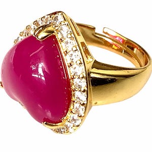 Peut inclure: Bague réglable dorée avec une pierre en forme de cœur, brillante, de couleur magenta. La pierre est encadrée par une monture dorée avec une bordure de petits cristaux clairs et étincelants. Le corps de la bague est également doré.