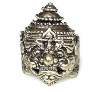Op de afbeelding: Een zilveren ring met een gedetailleerd ontwerp van een mythisch wezens gezicht. Het wezen heeft een grote, puntige neus, brede ogen en een woeste uitdrukking. De ring is versierd met ingewikkelde details en een kroonachtige structuur bovenop.