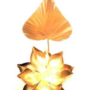 May include: A golden metal lotus flower with a large, heart-shaped leaf rising above it. The flower has multiple layers of petals, and the leaf and flower have a textured, metallic finish. The background is plain white.