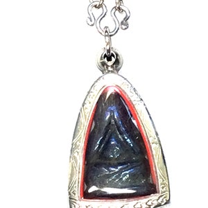 Puede incluir: Collar con colgante plateado con un marco triangular ornamentado. El colgante presenta un elemento central oscuro, posiblemente negro, enmarcado por un borde rojo. La cadena del collar también es plateada.