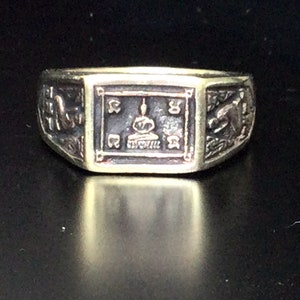 May include: A gold ring with a square design featuring a Buddha figure in the center. The ring has intricate details and a dark brown finish.