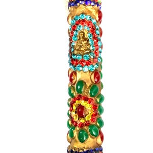 May include: A gold-colored, cylindrical decorative item adorned with colorful gemstones. The item features a central design with a small gold Buddha figure surrounded by red and blue gems. Green, red, and yellow gems are also incorporated.
