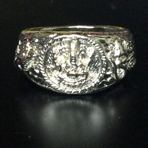 May include: A silver-colored ring with a detailed design. The ring features a central emblem surrounded by a textured border. The ring is set against a dark background, highlighting its intricate details and metallic sheen.