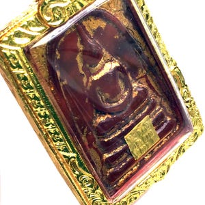 Puede incluir: Un colgante chapado en oro con un amuleto de Buda rojo y dorado en su interior. El amuleto está encerrado en una cubierta de plástico transparente. El colgante tiene un marco dorado detallado con diseños intrincados.