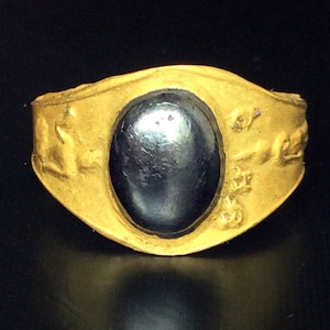 Puede incluir: Un anillo dorado con una piedra oscura de forma ovalada en el centro. La banda del anillo tiene un diseño texturizado, y la piedra parece pulida. El anillo se encuentra sobre un fondo oscuro, resaltando sus detalles.