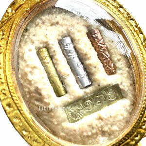 May include: A gold-colored pendant with a clear, oval-shaped center. Inside, there are three small, cylindrical metal pieces in gold, silver, and copper colors, along with a rectangular piece. The text "Jubilee Store" is visible.