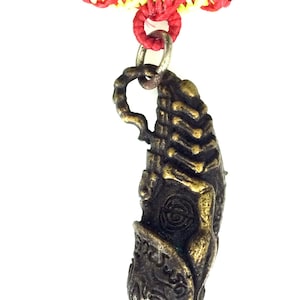 Può includere: Un ciondolo dorato a forma di mano con dettagli intricati, appeso a un cordoncino nero con accenti intrecciati rossi e gialli.