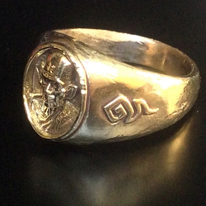 Op de afbeelding: Een gouden ring met een gedetailleerd ontwerp van een gezicht aan de voorkant en een gestileerd symbool aan de zijkant.