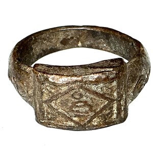 Op de afbeelding: Een verweerde, zilverkleurige ring met een rechthoekige voorkant. De voorkant heeft een ruitpatroon met een geëtst ontwerp in het midden. De ring heeft een versleten, antieke uitstraling, wat duidt op leeftijd en gebruik.