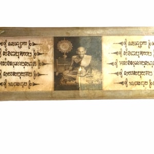 Puede incluir: Amuleto religioso rectangular con una imagen central en blanco y negro de un monje sentado. El amuleto tiene texto en un idioma desconocido a ambos lados de la imagen. El amuleto está encerrado en un marco de metal color plata.