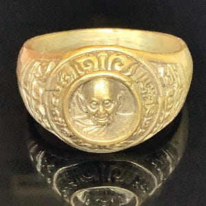 Op de afbeelding: Een goudkleurige ring met een reliëf ontwerp. De ring heeft een centraal medaillon met een gedetailleerde afbeelding van een gezicht. De band van de ring is versierd met ingewikkelde patronen. De ring wordt gepresenteerd op een reflecterend oppervlak.