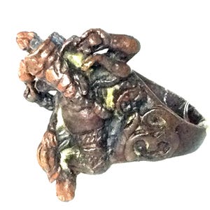 May include: A detailed bronze-colored ring featuring a sculpted figure with intricate details. The ring has a textured surface and a decorative design on the band, showcasing a unique, artistic style.