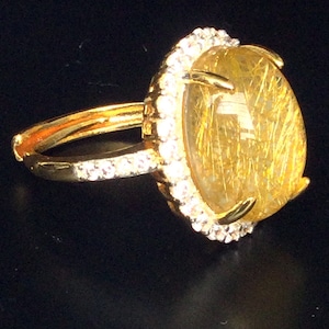 May include: A gold ring with a large, oval-cut, golden gemstone with internal golden inclusions, encircled by a halo of small, clear gemstones. The ring band is gold and set with small, clear gemstones.
