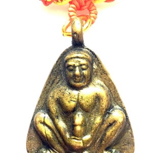 Puede incluir: Un colgante de color dorado que representa una figura sentada con un rostro y un cuerpo detallados. El colgante está unido a un cordón negro con detalles trenzados de color naranja y amarillo.