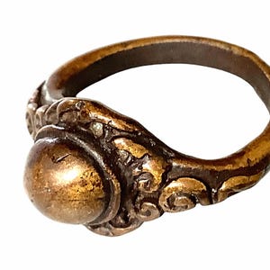 Puede incluir: Un anillo de bronce con un diseño ornamentado. El anillo presenta una piedra central grande y redondeada y un trabajo de volutas detallado en la banda. El metal tiene un aspecto envejecido, lo que sugiere un estilo antiguo o vintage.