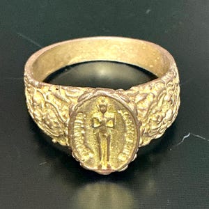Op de afbeelding: Een goudkleurige ring met een reliëf ontwerp. De ring heeft een centraal medaillon met een figuur in een lauwerkrans. De band heeft een gestructureerd, sierlijk patroon. De ring is waarschijnlijk van metaal.