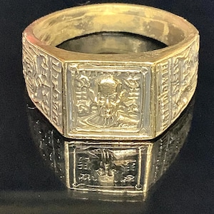 Op de afbeelding: Een goudkleurige ring met een rechthoekige voorkant met een gedetailleerd reliëf. De band van de ring heeft gegraveerde patronen en tekst. De ring wordt gepresenteerd op een reflecterend oppervlak.