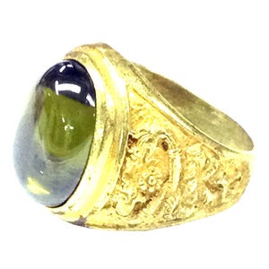 Puede incluir: Un anillo de oro con una piedra preciosa verde ovalada engastada en el centro. La banda del anillo está decorada con intrincados tallados.