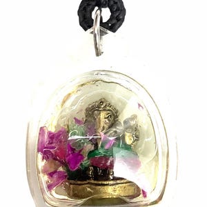 May include: A small, clear pendant featuring a gold-colored statue of a deity, possibly Ganesha, with purple flowers inside. The pendant hangs from a black and red braided cord.