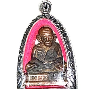 Puede incluir: Un colgante metálico que presenta una figura de bronce de un monje sentado, encerrado en un marco de color plateado con intrincados patrones de volutas. La figura se sitúa sobre un fondo rosa vibrante y la base incluye escritura tailandesa.