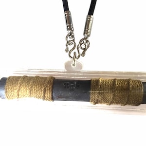 Könnte beinhalten: Ein rechteckiger Anhänger mit einem dunklen zylindrischen Kern, der mit beigefarbenem Bindfaden umwickelt ist und in einem transparenten Kunststoffgehäuse eingeschlossen ist. Der Anhänger hängt an einer schwarzen Kordel mit silberfarbenen Metallverschlüssen. Der Anhänger hat Text.