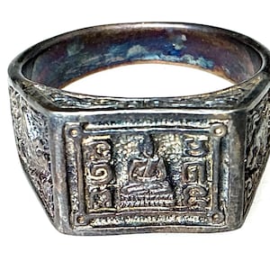 May include: A silver ring with a rectangular face, displaying a seated figure and detailed ornamentation. The ring has an aged look, with intricate patterns on the sides and a slightly tarnished finish.
